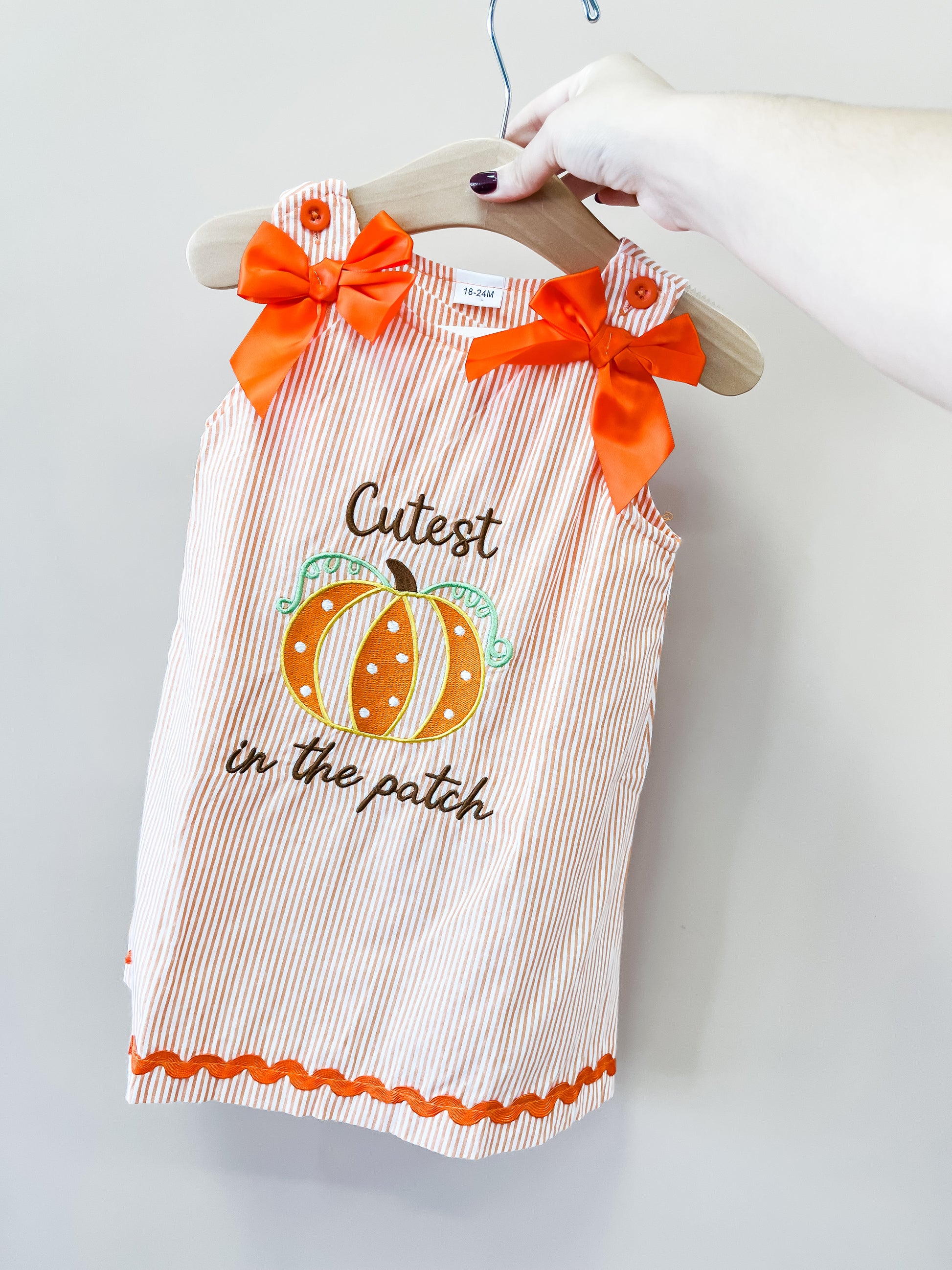 A hand holds a hanger with the Tiny Tot Bundles “Cutest In The Patch Dress,” a striped orange-and-white toddler dress featuring orange bows and an adorable pumpkin graphic, with the words “Cutest in the patch” on the front.