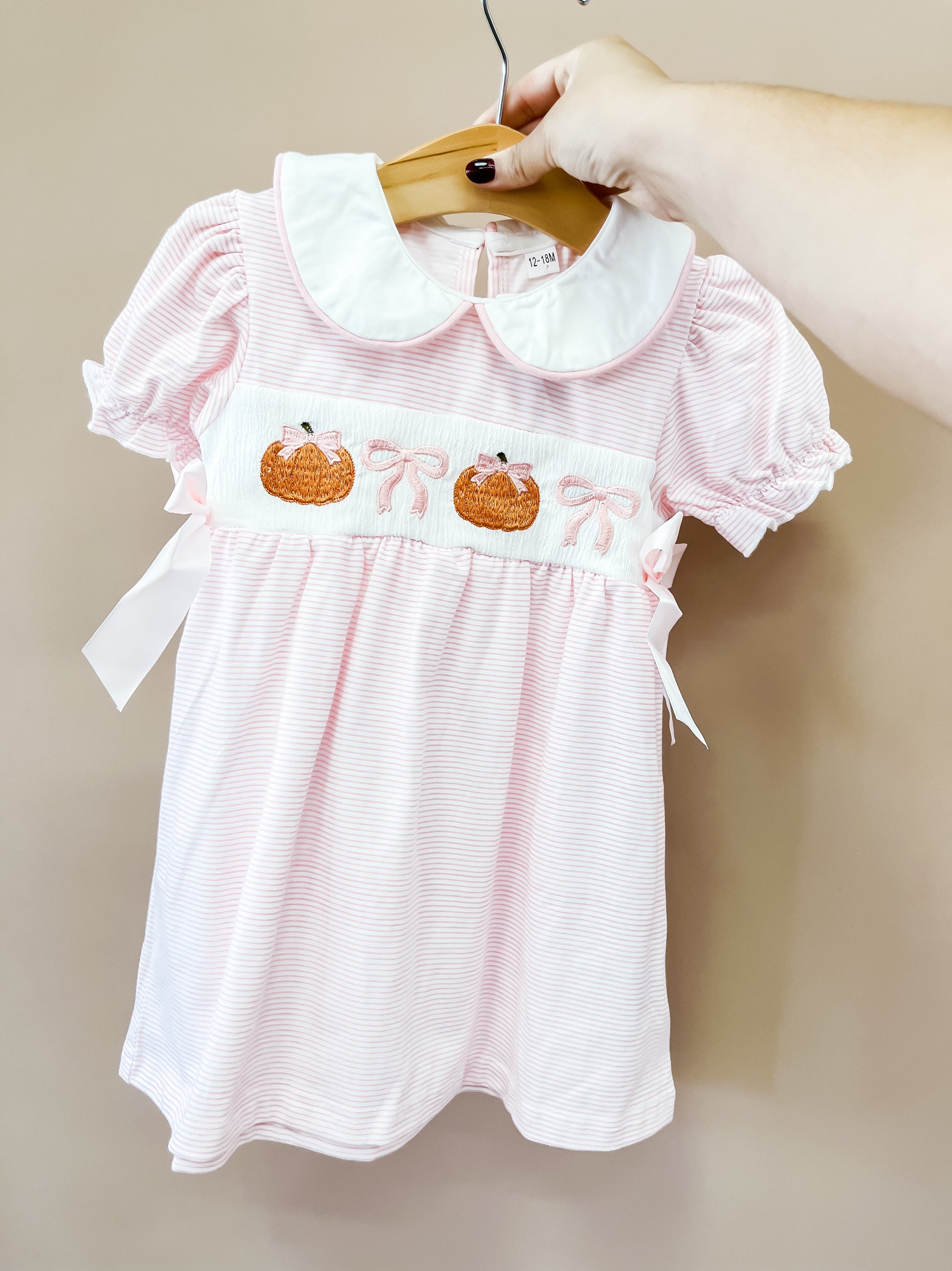 A hand holds up the Tiny Tot Bundles Pink Pumpkin Bow Dress, featuring pink and white stripes, puff sleeves, a white collar, pink bows, and pumpkin embroidery on the front.
