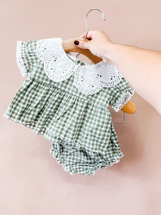 A hand holds the Tiny Tot Bundles Evergreen Gingham Set—featuring a green and white checkered baby dress with a white scalloped collar and matching bloomers—on a wooden hanger, set against a light pink background.