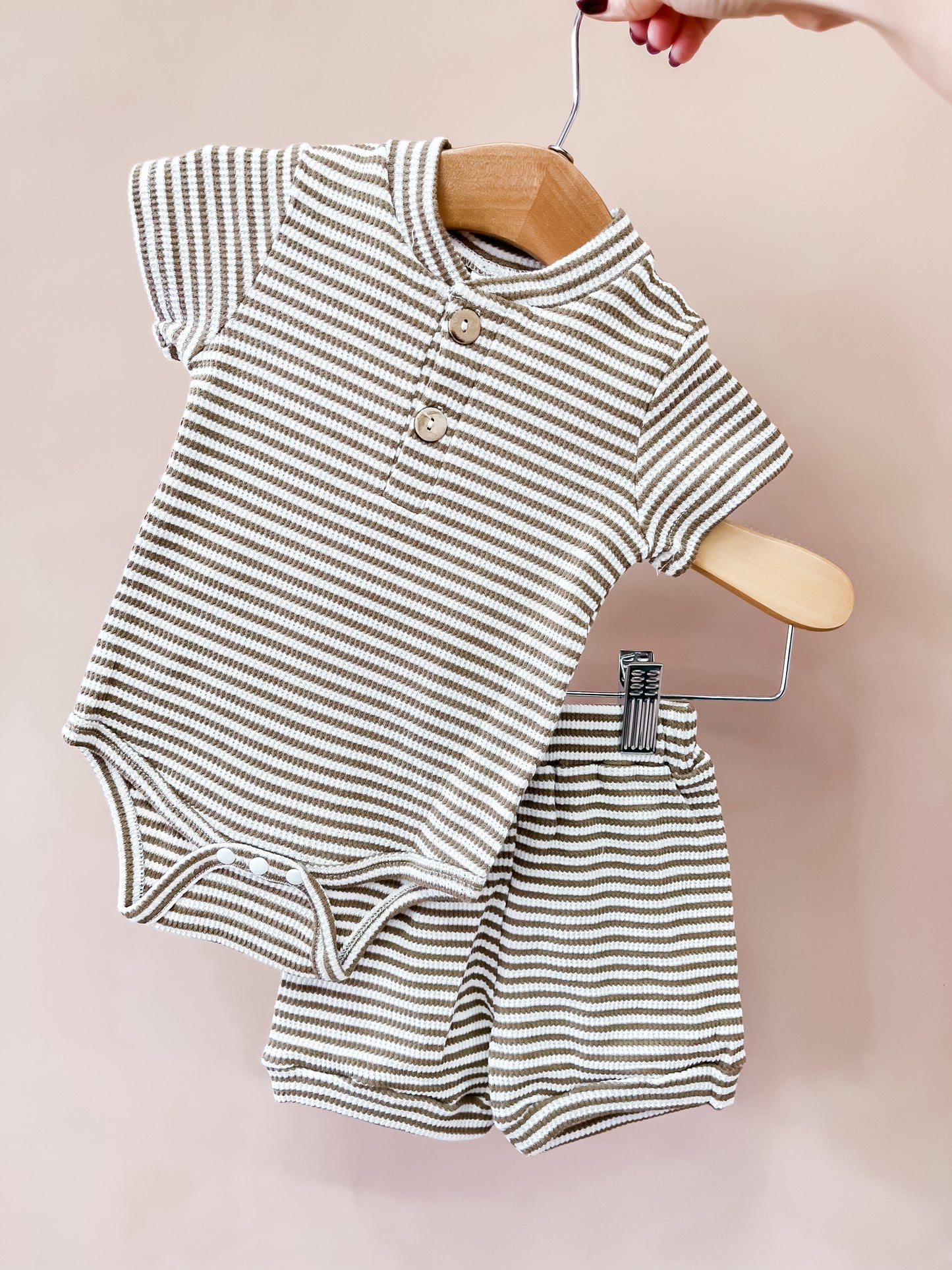 A hand holds a hanger featuring the Tiny Tot Bundles Harvest Stripe Infant Set, which includes a beige and white striped baby onesie and matching shorts, set against a plain light pink background.
