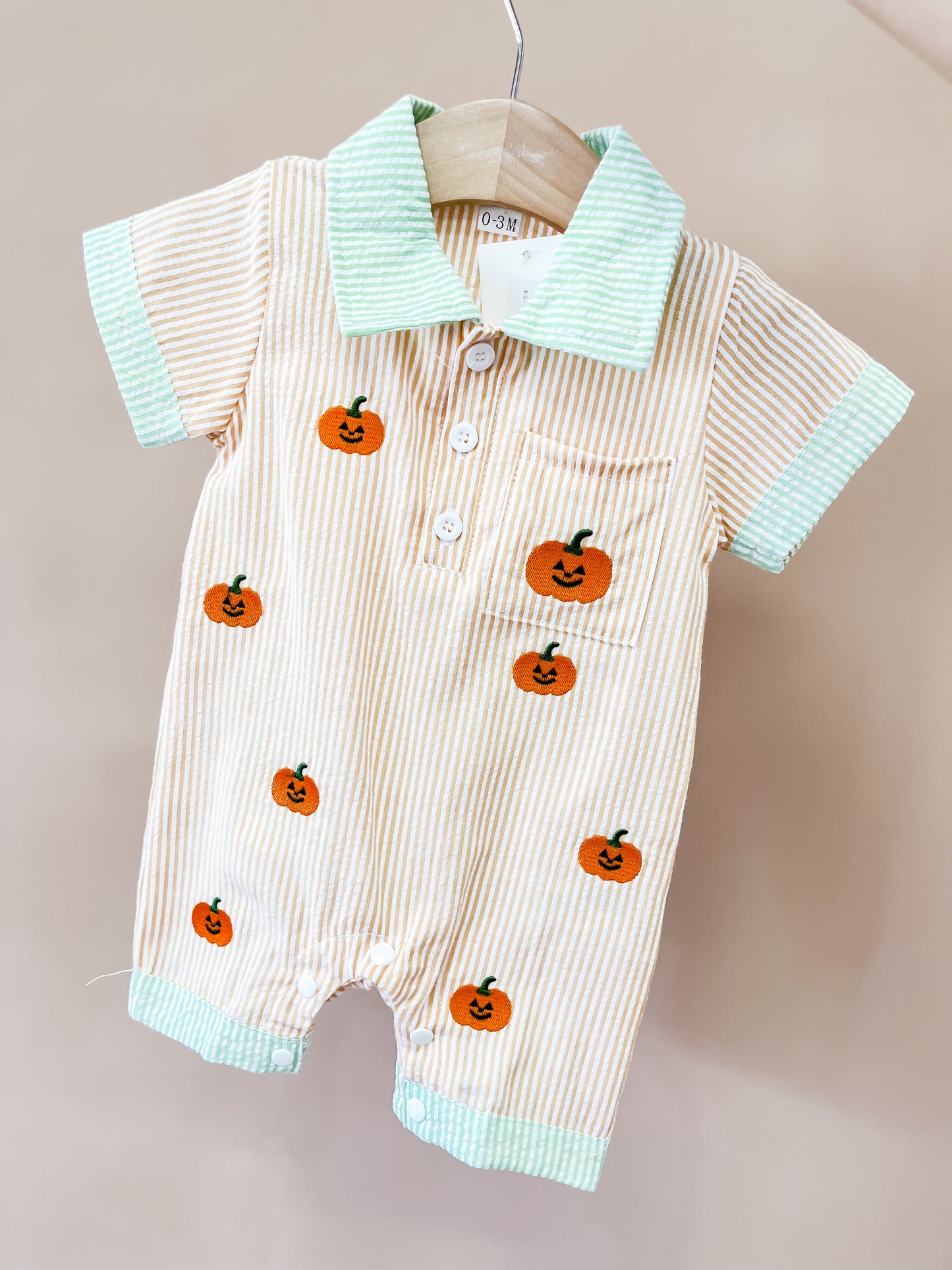 The Pumpkin Parade Collard Romper from Tiny Tot Bundles features thin beige stripes with green trim and is adorned with cheerful, smiling orange pumpkins. This short-sleeved romper comes with a collar, front buttons, and a handy chest pocket.