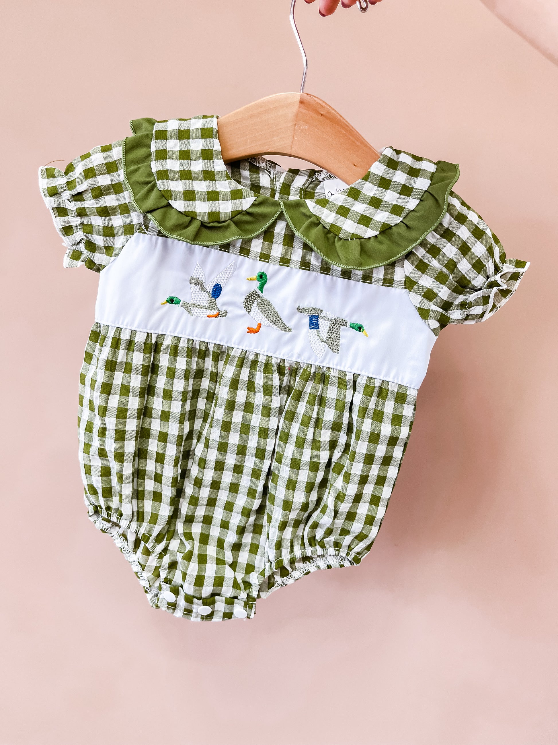 The Mallard Meadow Bubble by Tiny Tot Bundles is a green and white gingham baby romper featuring a charming Peter Pan collar, short sleeves, and embroidered ducks across a white chest panel. Perfect for fall 2026, the romper is displayed on a wooden hanger against a light pink background.