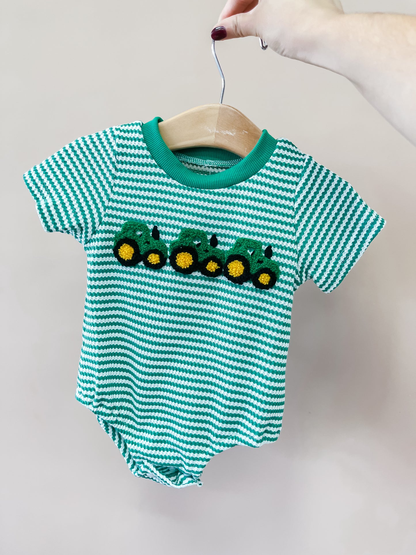 A hand holds the Tractor Trails Green Romper by Tiny Tot Bundles—a green and white striped baby romper on a hanger, featuring three green tractors across the chest.