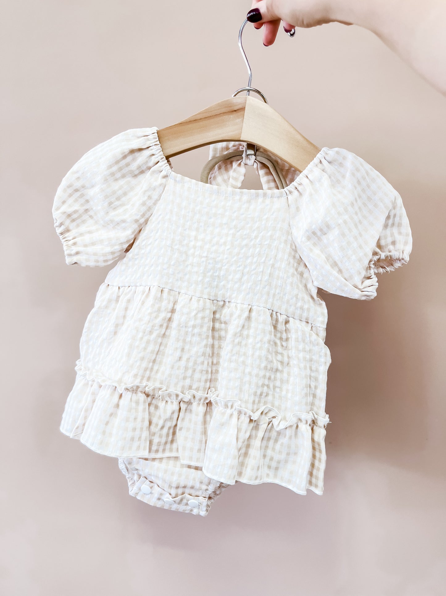A hand holds a hanger with the Tiny Tot Bundles Honey Oat Gingham Romper, featuring cream and beige checkered fabric, puffed short sleeves, and a ruffled skirt, displayed against a plain beige background.