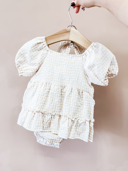 A hand holds a hanger with the Tiny Tot Bundles Honey Oat Gingham Romper, featuring cream and beige checkered fabric, puffed short sleeves, and a ruffled skirt, displayed against a plain beige background.