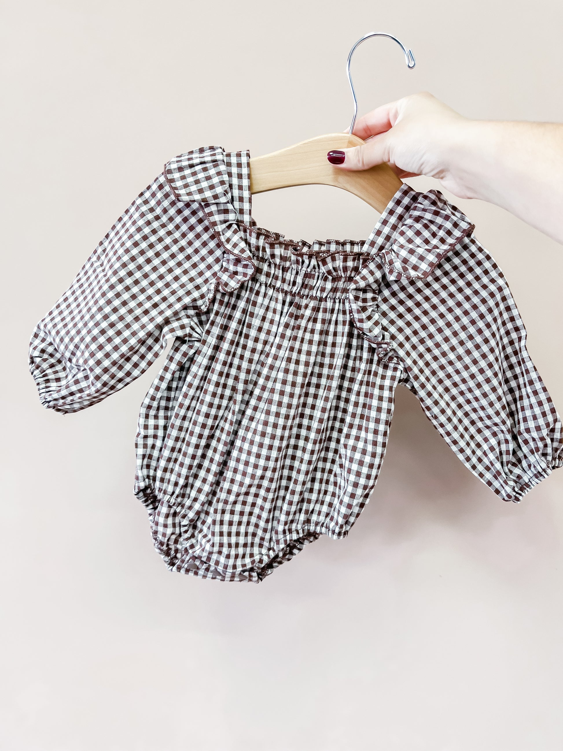 A hand holds a hanger displaying the Tiny Tot Bundles Autumn Gingham Bubble, a long-sleeved baby romper featuring a brown and white gingham pattern, set against a plain light background.