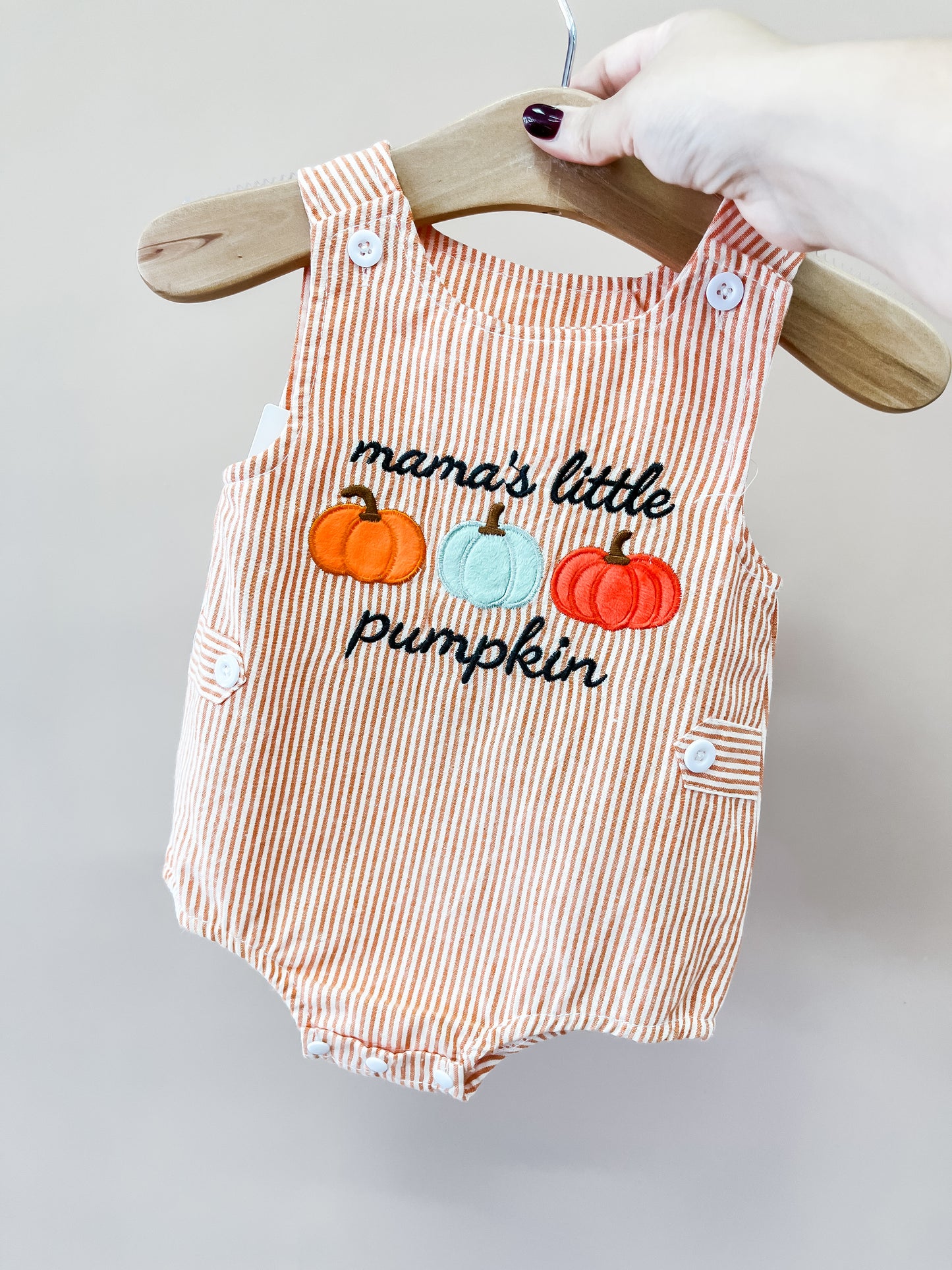 A hand holds the Tiny Tot Bundles Mama’s Pumpkin Stripe Romper on a hanger. The romper features orange and white stripes with embroidered pumpkins and “mama’s little pumpkin” on the front.