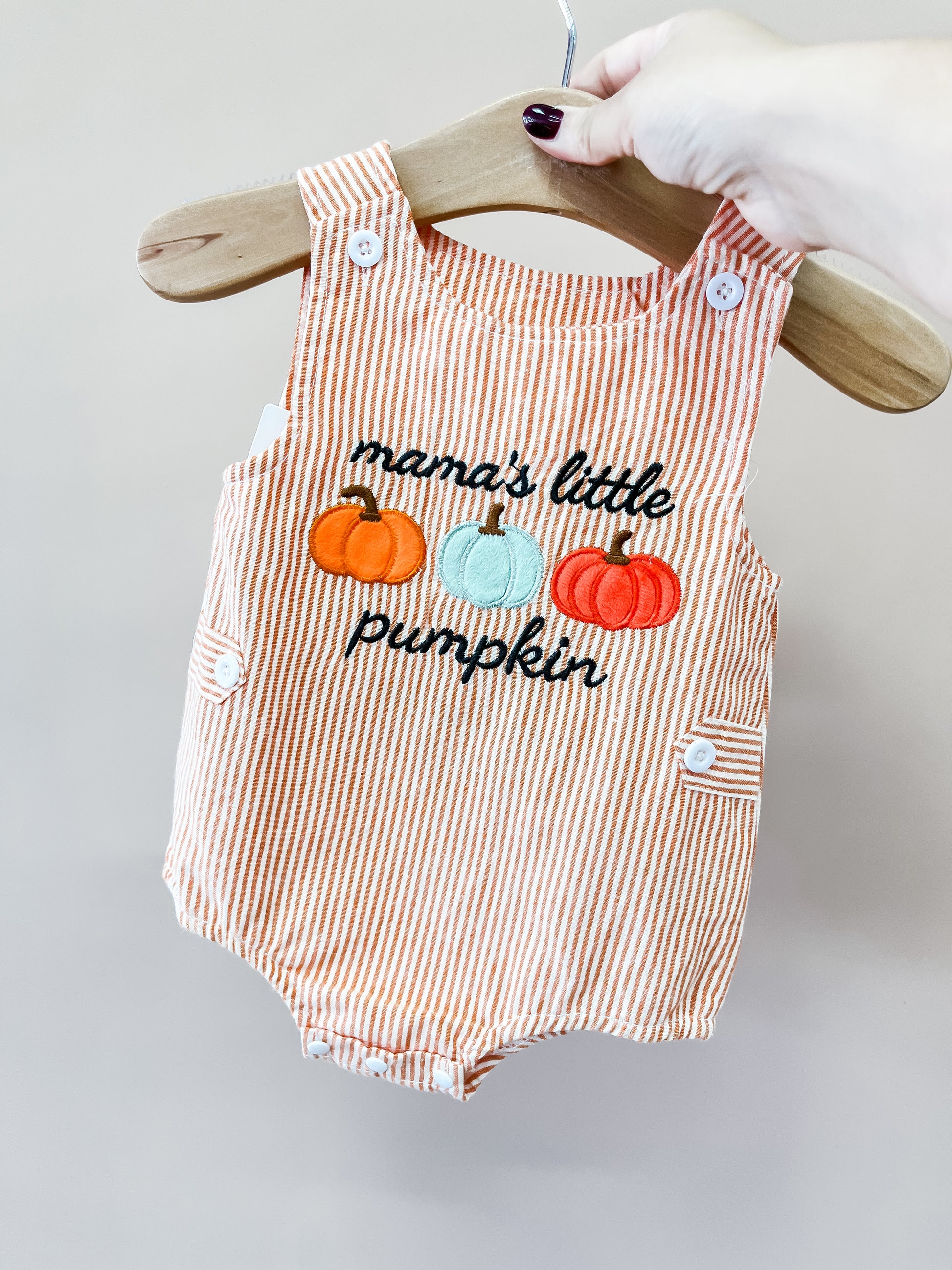 A hand holds the Tiny Tot Bundles Mama’s Pumpkin Stripe Romper on a hanger. The romper features orange and white stripes with embroidered pumpkins and “mama’s little pumpkin” on the front.