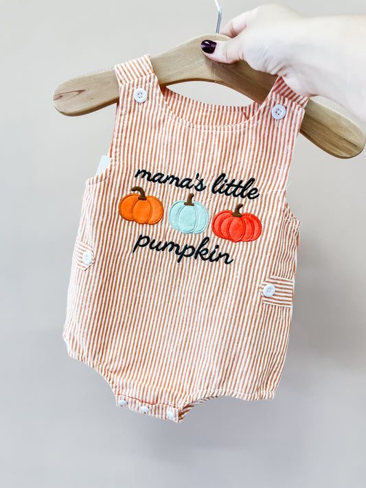 A hand holds the Tiny Tot Bundles Mama’s Pumpkin Stripe Romper on a hanger. The romper features orange and white stripes with embroidered pumpkins and “mama’s little pumpkin” on the front.