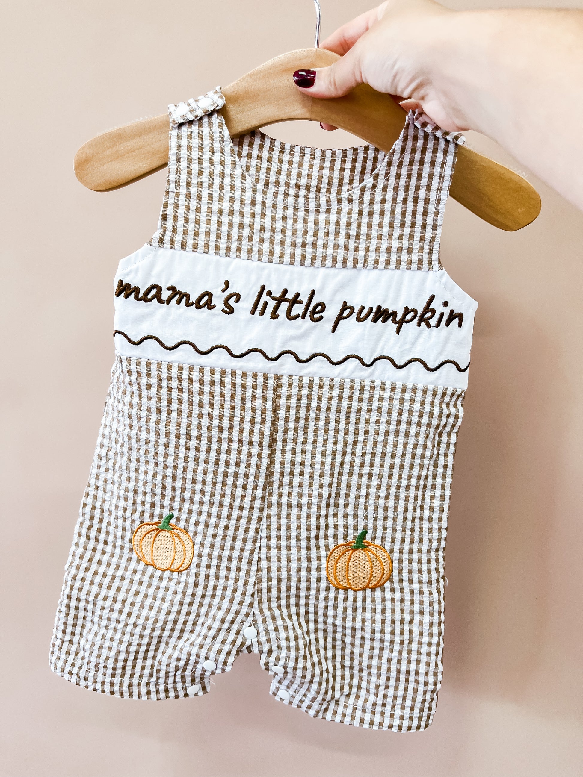 A hand holds the Mama’s Little Pumpkin Romper by Tiny Tot Bundles, a brown-and-white gingham baby romper on a hanger, featuring embroidered pumpkins and the words “Mama’s Little Pumpkin” on the chest.