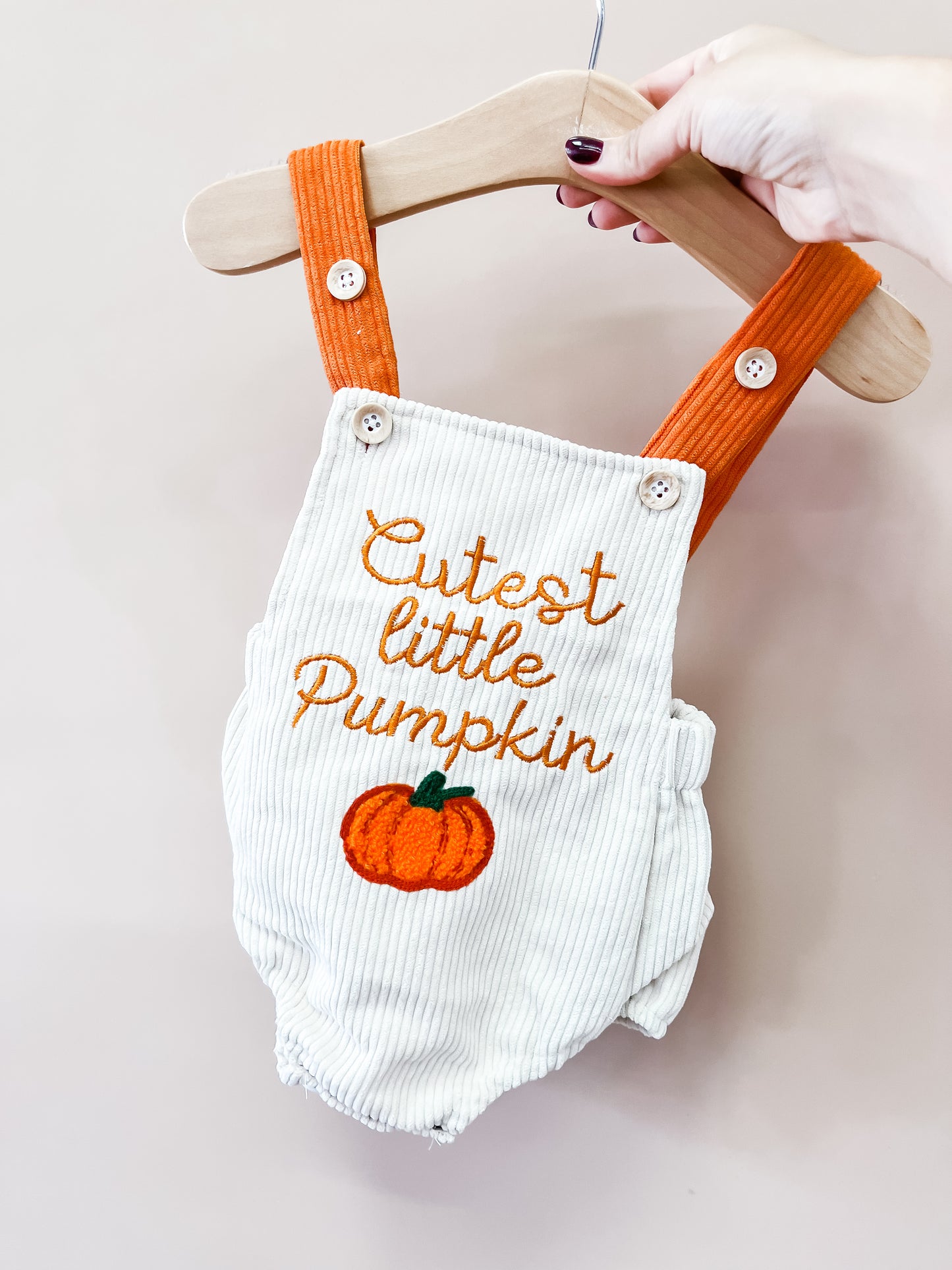 The Little Pumpkin Corduroy Overalls by Tiny Tot Bundles feature a cream corduroy design with orange straps, embroidered “Cutest little Pumpkin” text, and a pumpkin motif. They are displayed on a wooden hanger held by a hand with painted nails.