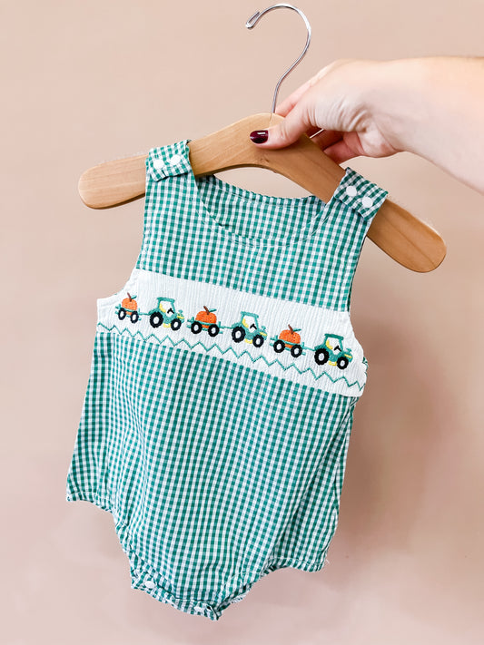 A hand holds the Tiny Tot Bundles Harvest Hayride Romper—a green and white gingham baby romper with embroidered colorful tractors across the chest—on a wooden hanger.