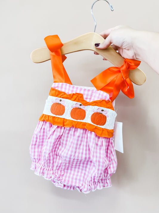 A hand holds the Little Miss Pumpkin Romper by Tiny Tot Bundles—a pink gingham baby romper featuring orange ribbons and embroidered pumpkins on the front—displayed on a wooden hanger against a light beige background.