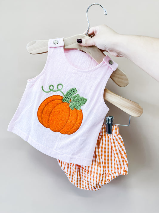 A hand holds a hanger with Tiny Tot Bundles’ Blushing Pumpkin Set—a pink toddler tank top adorned with a pumpkin patch and matching orange gingham shorts.