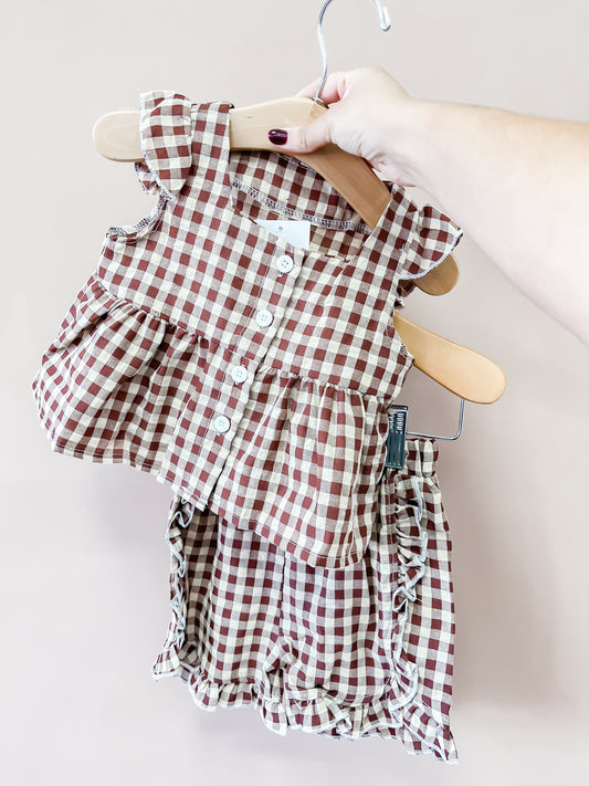 A hand holds a hanger displaying the Tiny Tot Bundles Cocoa Gingham Ruffle Set, featuring a red and white gingham sleeveless top with ruffles and buttons, paired with matching ruffled shorts, against a light beige background.