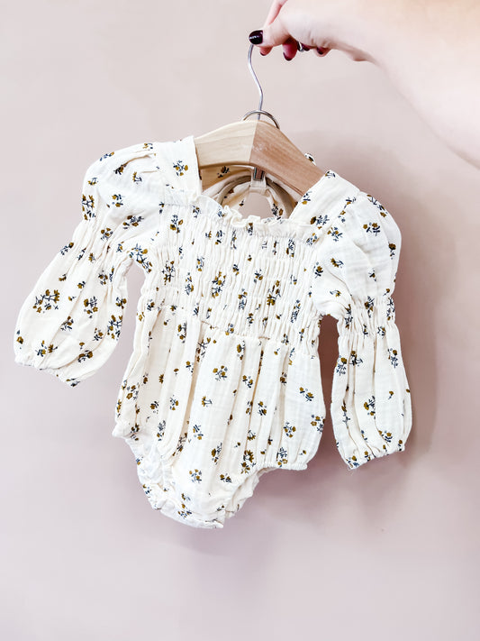 A hand holds a wooden hanger with the Golden Meadow Romper by Tiny Tot Bundles, a white, long-sleeve baby romper adorned with small floral prints and smocked detailing, set against a light pink background.