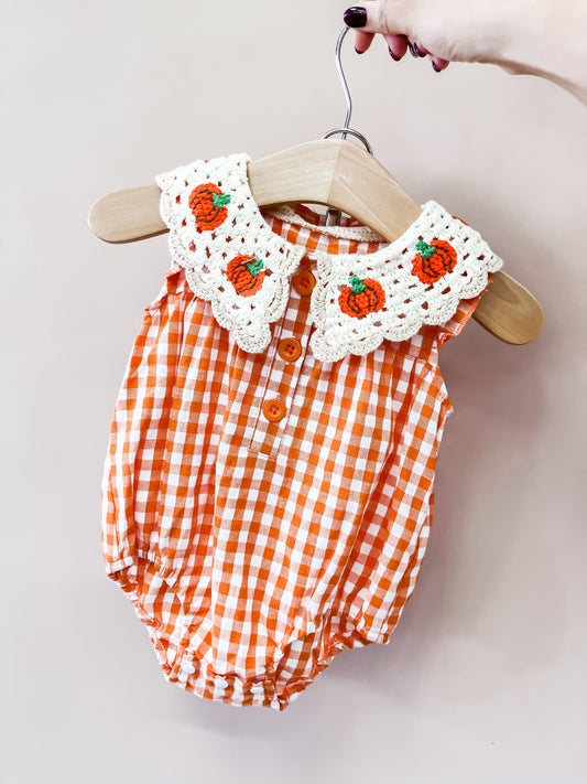 A hand holds a hanger displaying the Tiny Tot Bundles Vintage Gingham Romper—an orange-and-white checkered baby outfit with a white crochet collar adorned with small orange pumpkin designs.