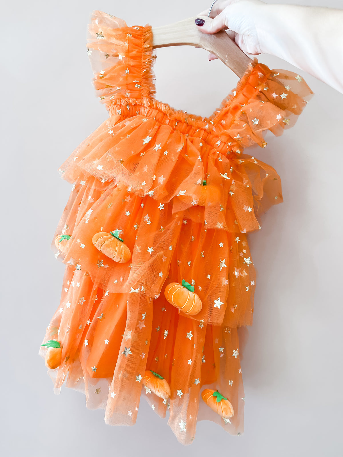 A hand with painted nails holds the Tiny Tot Bundles “Pumpkin Patch Tulle Dress,” an orange, ruffled child’s dress featuring gold stars and small pumpkin decorations.