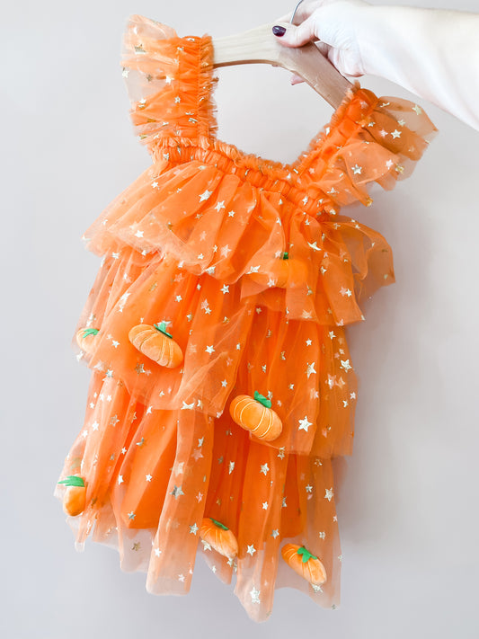 A hand with painted nails holds the Tiny Tot Bundles “Pumpkin Patch Tulle Dress,” an orange, ruffled child’s dress featuring gold stars and small pumpkin decorations.