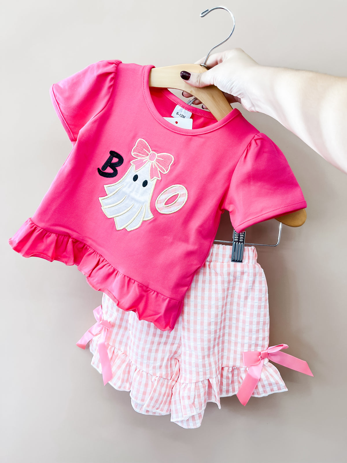 A hand holds the Tiny Tot Bundles Pink Boo Ghost Gingham Set, which includes a ruffled ghost shirt and matching pink gingham shorts with bows and ruffled hems, against a plain background.