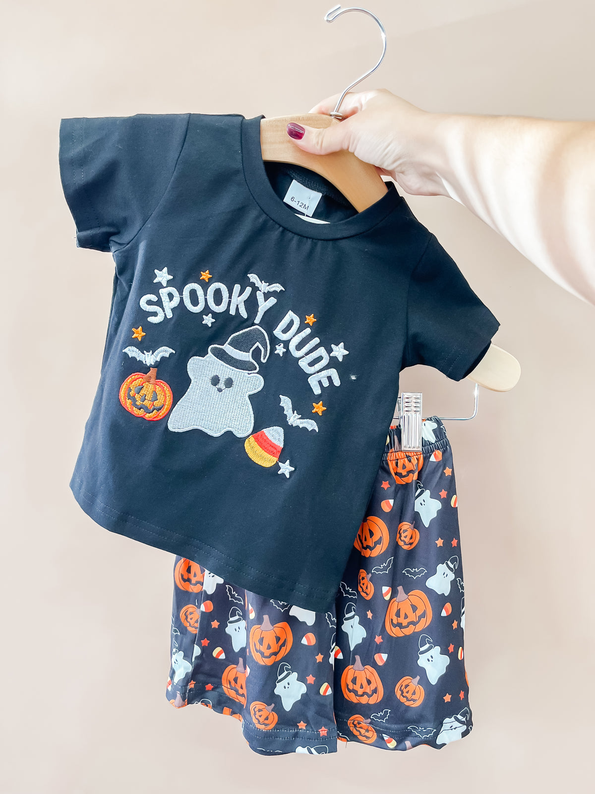 A hand holds up the Tiny Tot Bundles Spooky Dude Halloween Set, which includes a black shirt with a ghost and “Spooky Dude” text, paired with matching shorts decorated with ghosts, pumpkins, and stars.