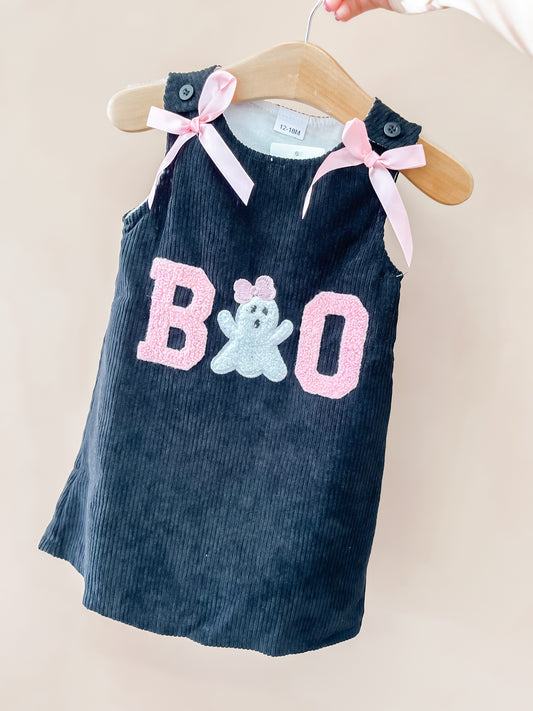 The Spooky Boo Dress by Tiny Tot Bundles is a dark corduroy toddler dress displayed on a hanger, featuring pink bows on the shoulders, “BOO” in pink letters, and a ghost applique on the front.