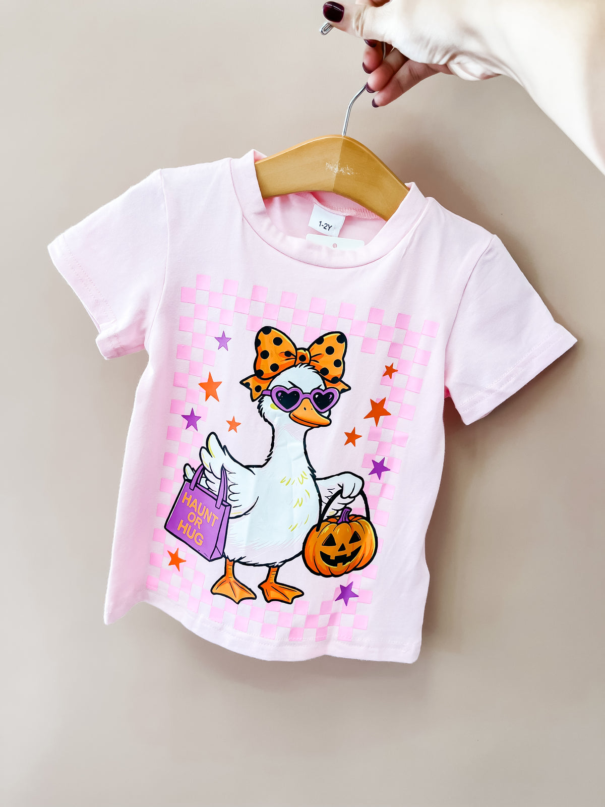 The Pink Spooky Goose Tee by Tiny Tot Bundles is a toddler T-shirt in pink, showcasing a cartoon duck with a bow, sunglasses, handbag, and pumpkin bucket. The shirt is displayed on a wooden hanger held by hand.
