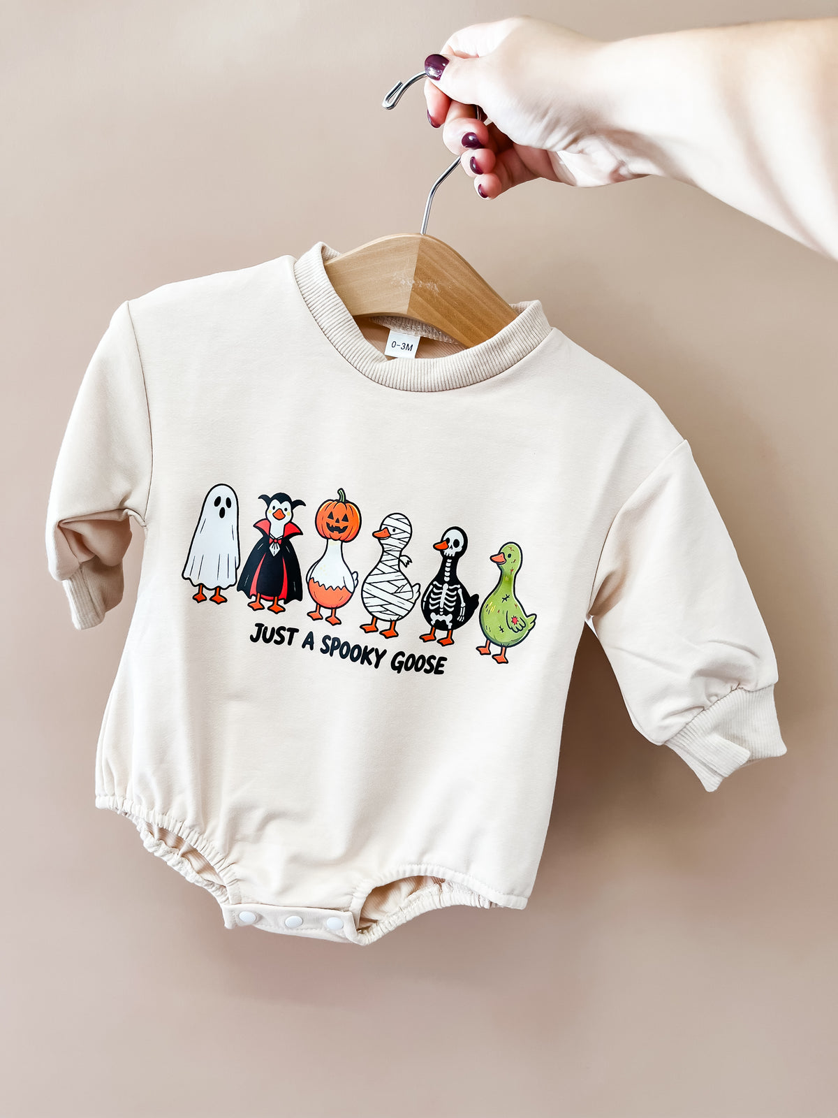 A hand holds the Tiny Tot Bundles “Just a Spooky Goose Romper” on a hanger. The romper displays five cartoon geese dressed in Halloween costumes—ghost, vampire, pumpkin, mummy, and skeleton—alongside one plain goose. The text reads: JUST A SPOOKY GOOSE.