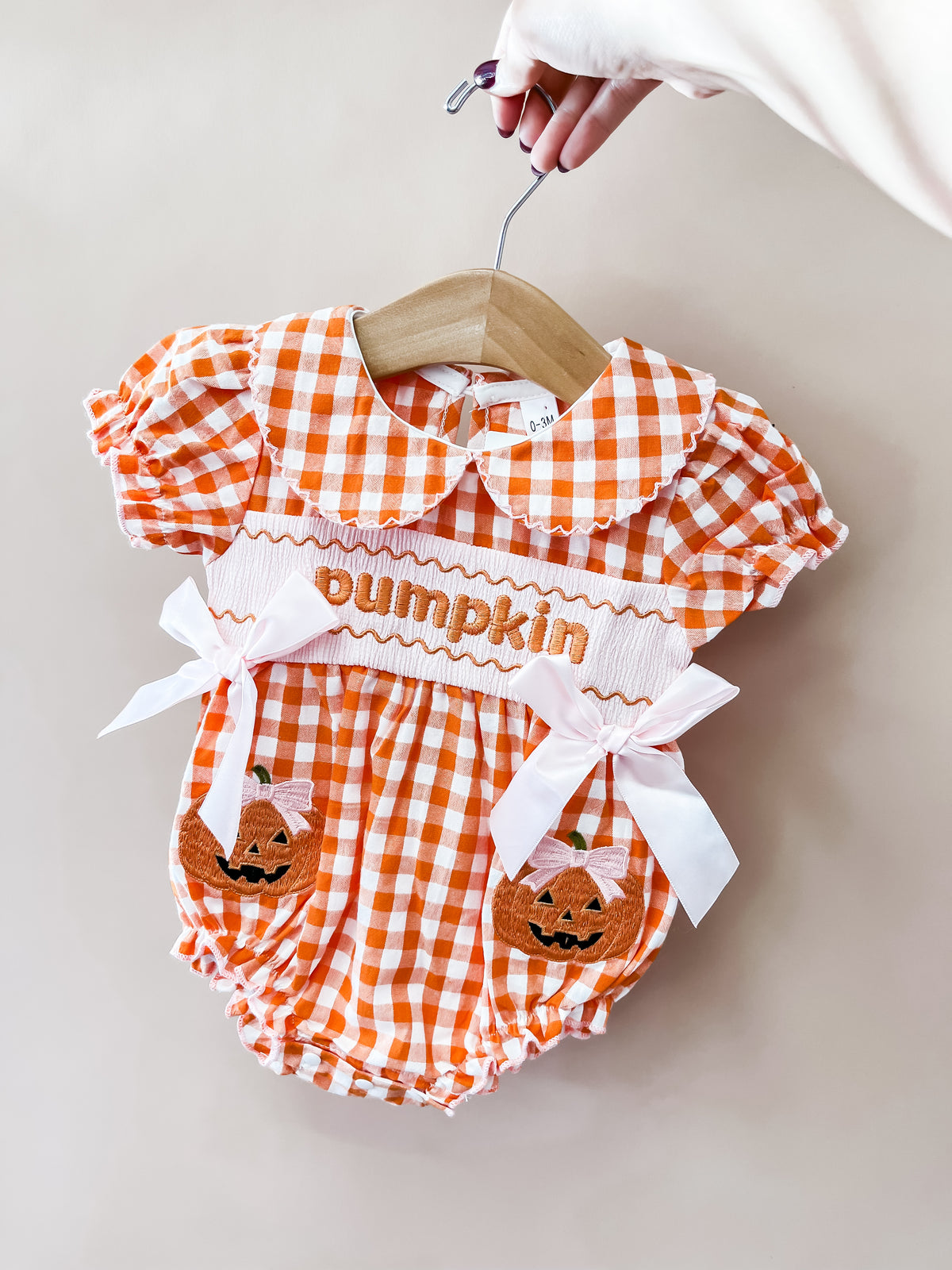 A hand holds a hanger with the Tiny Tot Bundles Orange Gingham Pumpkin Romper, which features “pumpkin” text, white bows, and embroidered jack-o-lanterns on the front.