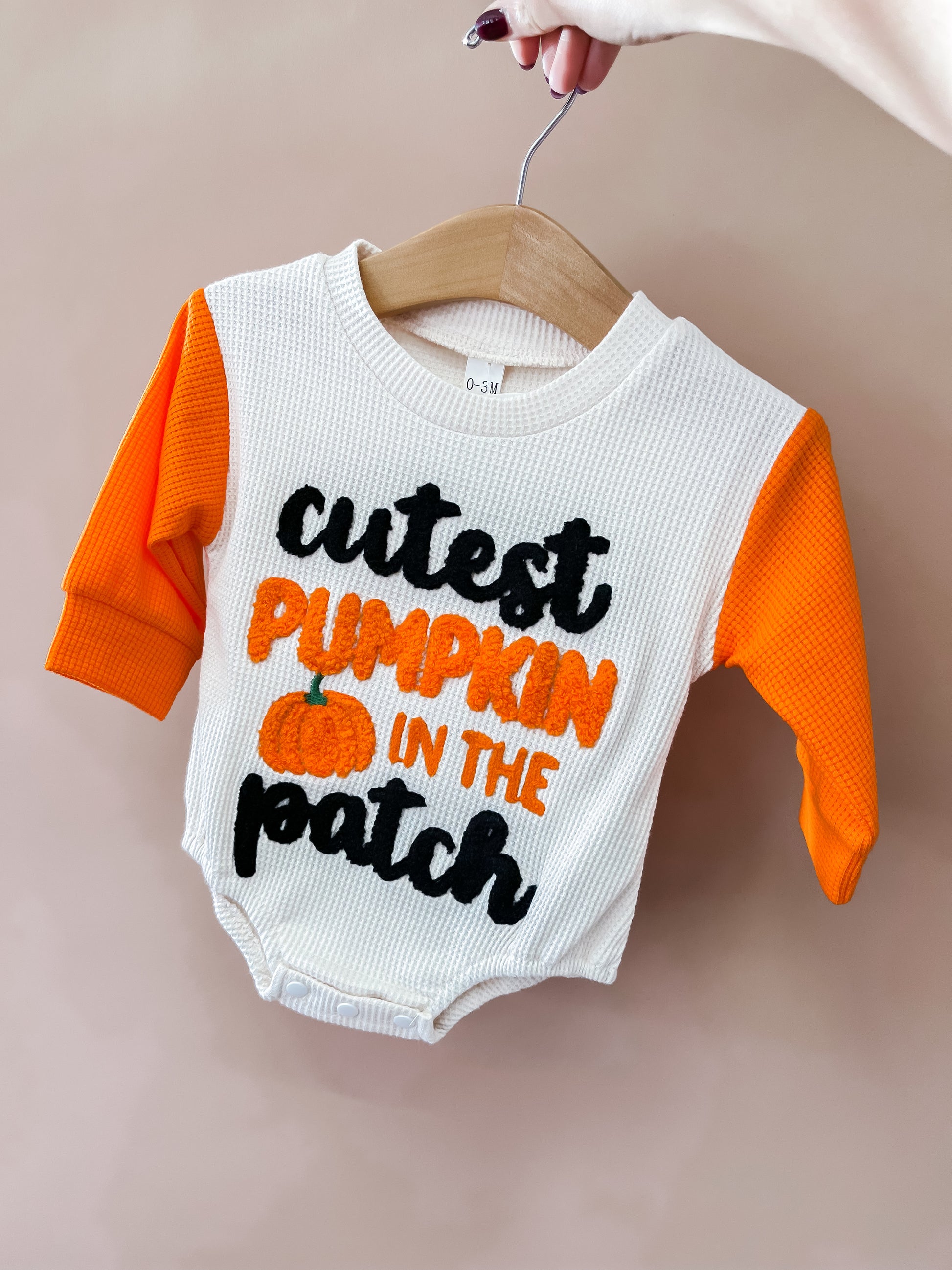 A hand holds the Tiny Tot Bundles Cutest Pumpkin in the Patch Romper on a hanger. The romper features orange sleeves and the phrase “cutest pumpkin in the patch” with a pumpkin illustration on the front.