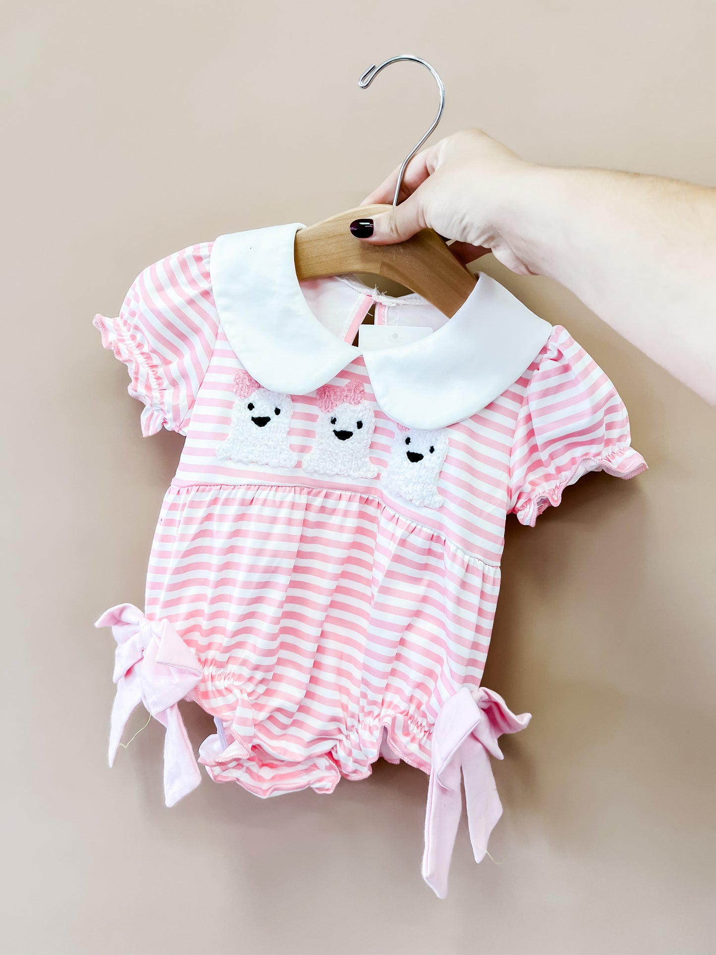 A hand holds the Tiny Tot Bundles Pink Stripe Ghost Romper on a hanger. The romper features pink and white stripes, a white Peter Pan collar, embroidered bear faces, and bow accents on the legs.