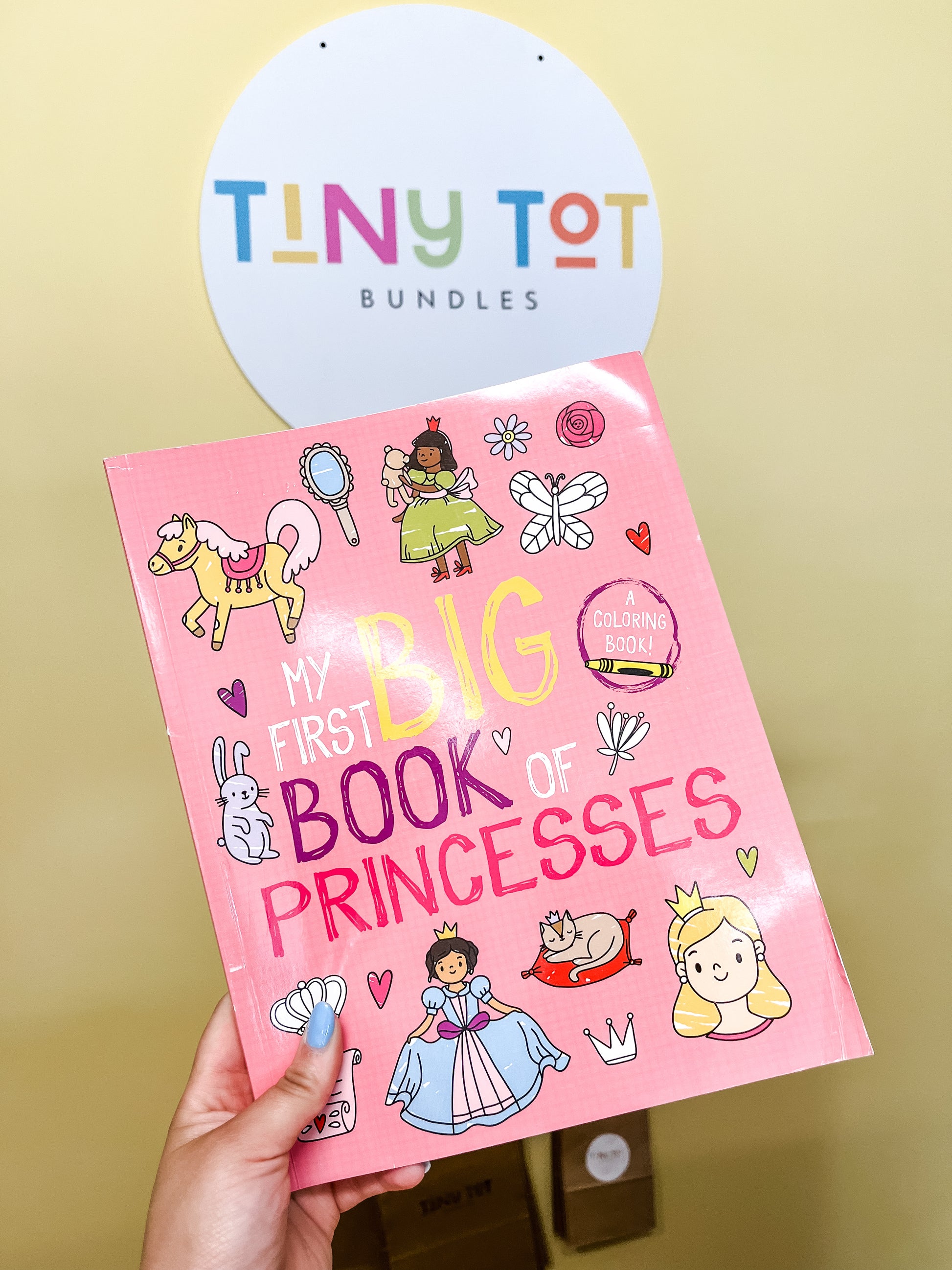 A hand holds the My First Big Book Coloring Book by Tiny Tot Bundles, featuring cartoon princesses and animals on the pink cover. A circular Tiny Tot Bundles sign is visible in the background.