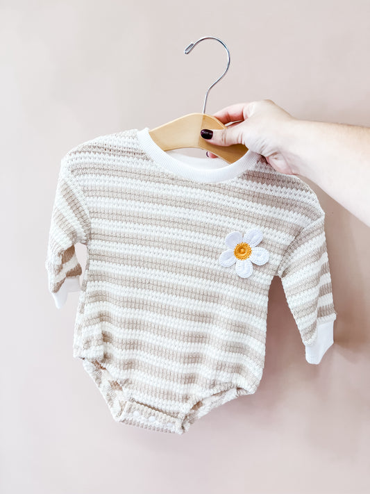 A hand holds the Tiny Tot Bundles Daisy Days Knit Romper, a beige and white striped baby outfit with long sleeves and a daisy flower patch on the chest, displayed on a wooden hanger against a pale background.
