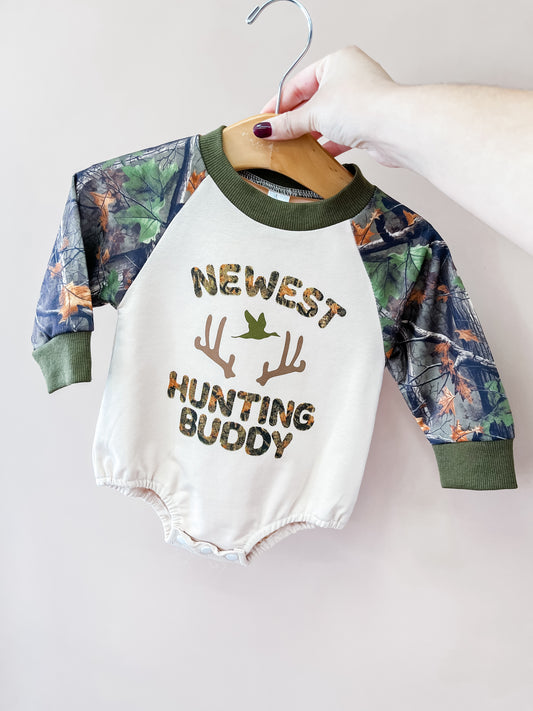 A hand holds the Tiny Tot Bundles Hunting Buddy Camo Romper, featuring camouflage sleeves, “Newest Hunting Buddy” text, antler graphics, and a bird illustration on the front.