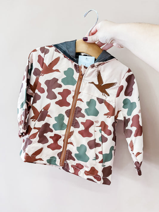 A hand holds the Tiny Tot Bundles Duck Hunter Windbraker on a hanger—a child’s jacket with a zipper and hood, featuring a brown, green, and beige camouflage pattern adorned with flying birds.