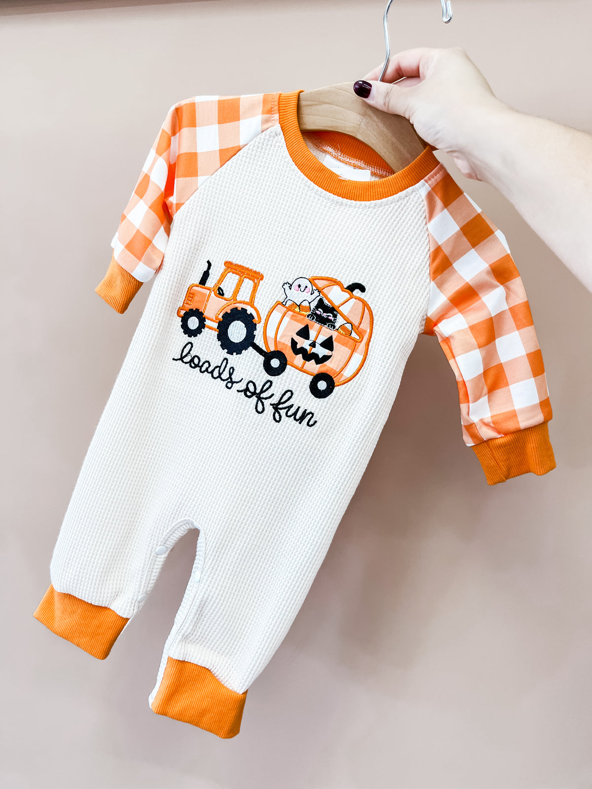 A hand holds the Trick Or tractor Romper by Tiny Tot Bundles, featuring orange plaid sleeves, cuffs, a tractor with a pumpkin, cat and ghost, and loads of fun embroidered on the front.