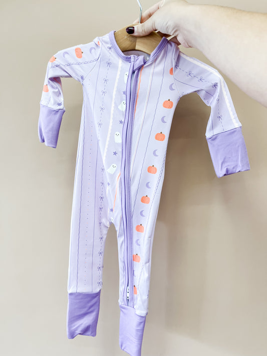 A hand holds up the Lavender Haunt Zippy by Tiny Tot Bundles, a lavender baby onesie with purple cuffs, a front zipper, and decorated with pumpkins, ghosts, moons, and stars.