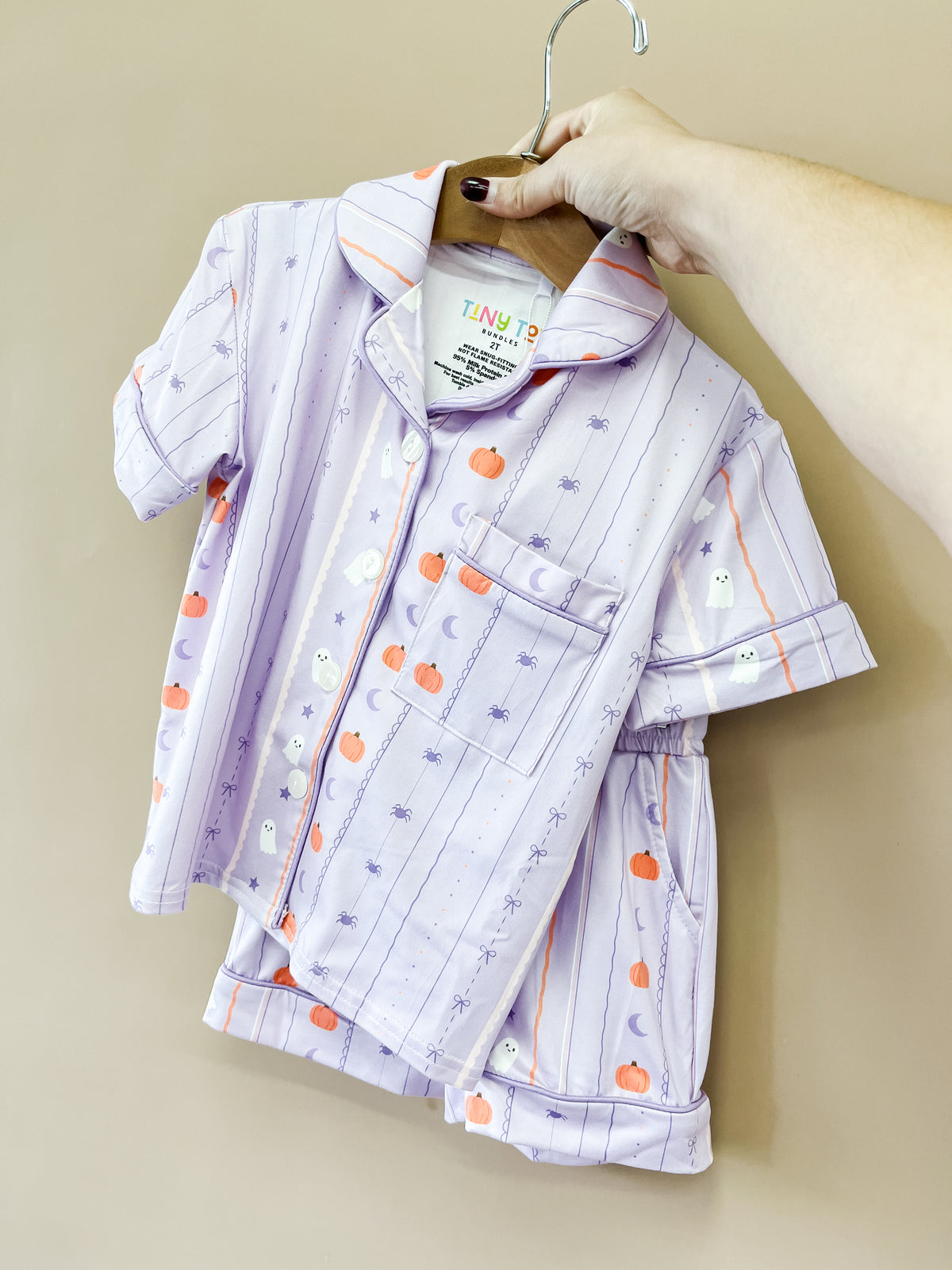 A hand holds up the Lavender Haunt Short Jammies Set by Tiny Tot Bundles, featuring pumpkins, ghosts, and bats on light purple short sleeves, displayed on a hanger against a beige background.