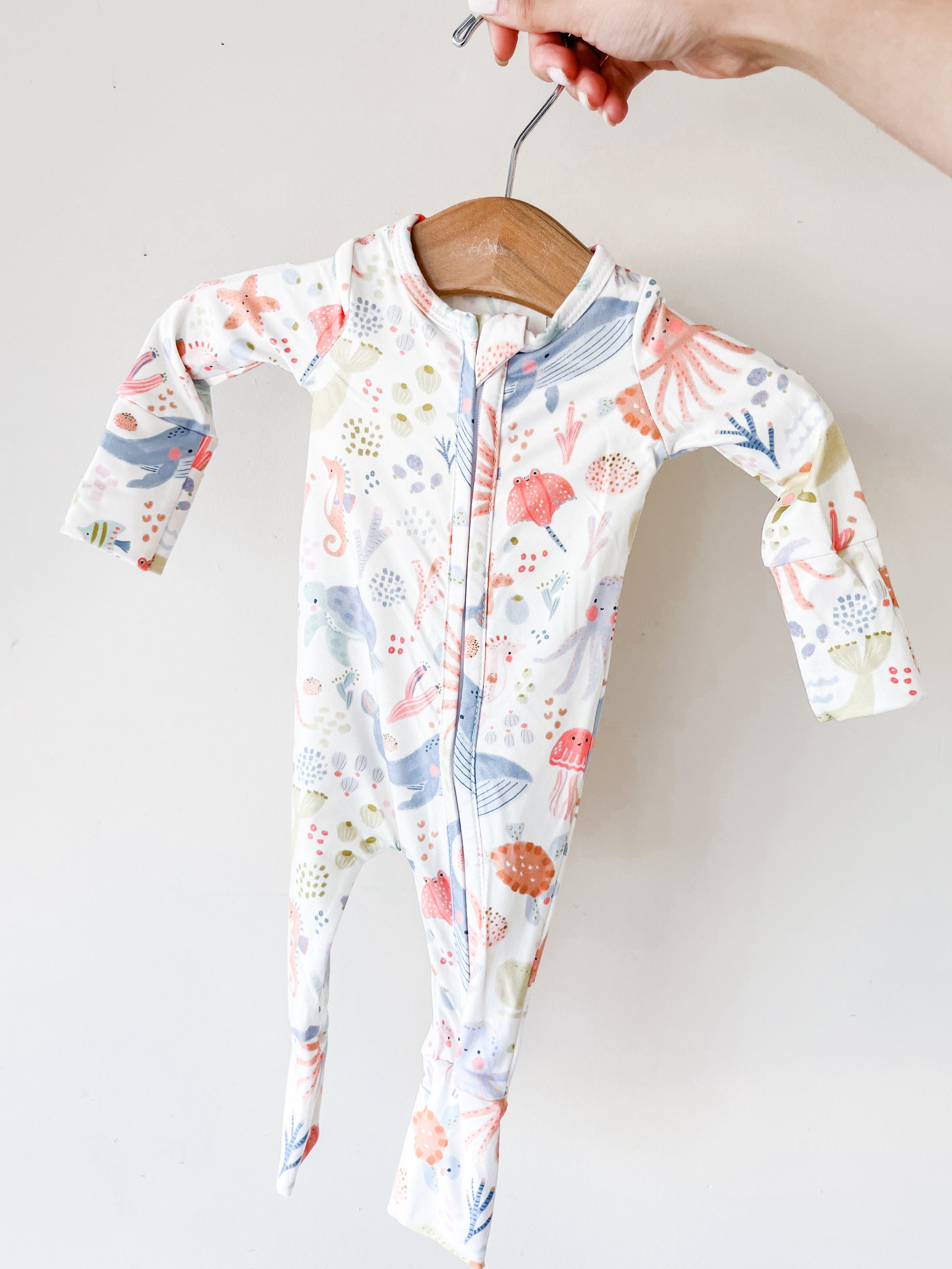 A hand holds the Tiny Tot Bundles Florida Reef Long Zippy, a colorful baby onesie with a whimsical underwater pattern of whales, fish, and coral, displayed on a wooden hanger against a plain white background.