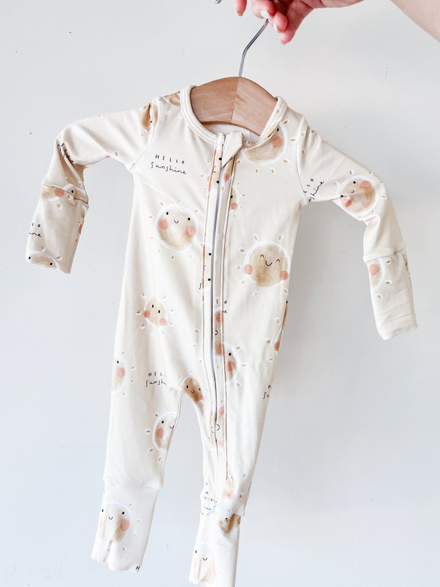 A hand holds the Tiny Tot Bundles Sunshine State Long Zippy, a cream baby onesie with sun and cloud patterns, smiling faces, and "Hello Sunshine" printed on it, set against a plain white background.
