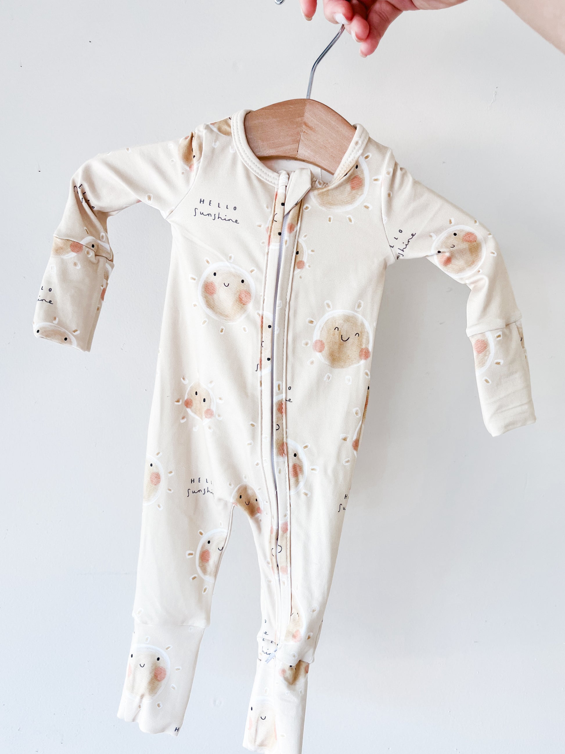 A hand holds the Tiny Tot Bundles Sunshine State Long Zippy, a cream baby onesie with sun and cloud patterns, smiling faces, and "Hello Sunshine" printed on it, set against a plain white background.