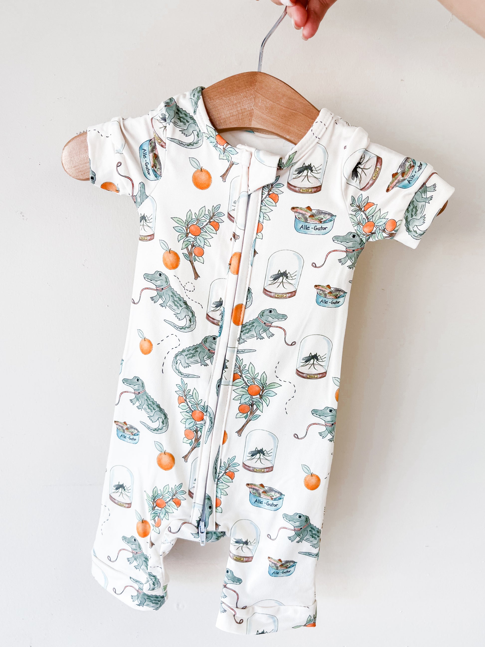 The Tiny Tot Bundles Sunshine Swamp Short Zippy, a short-sleeved baby romper with a zipper and alligator, orange, flower, and jar prints, hangs on a wooden hanger held by hand against a plain white background.
