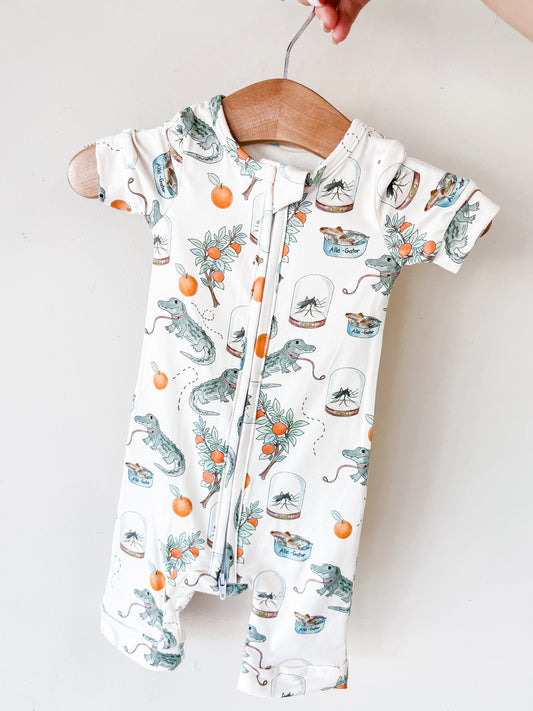The Tiny Tot Bundles Sunshine Swamp Short Zippy, a short-sleeved baby romper with a zipper and alligator, orange, flower, and jar prints, hangs on a wooden hanger held by hand against a plain white background.