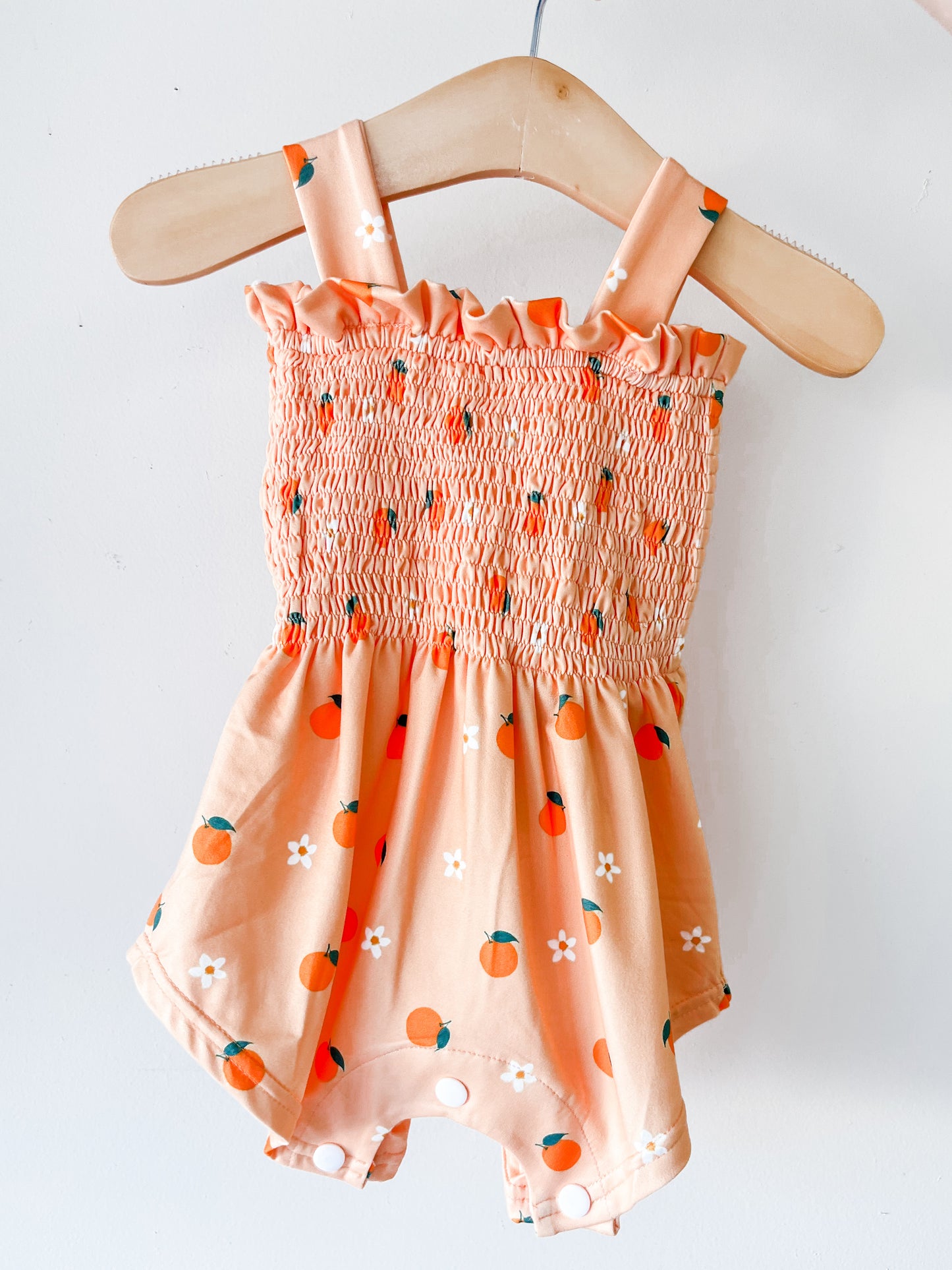 The Orange Blossom Smocked Romper by Tiny Tot Bundles features a peach color with orange fruit and white flower patterns, smocked bodice, ruffled neckline, and shoulder straps, displayed on a wooden hanger against a white background.