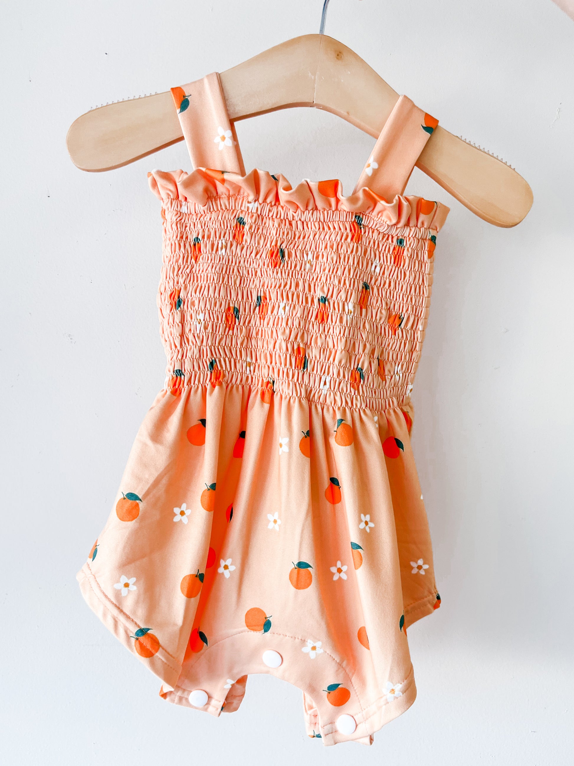 The Orange Blossom Smocked Romper by Tiny Tot Bundles features a peach color with orange fruit and white flower patterns, smocked bodice, ruffled neckline, and shoulder straps, displayed on a wooden hanger against a white background.