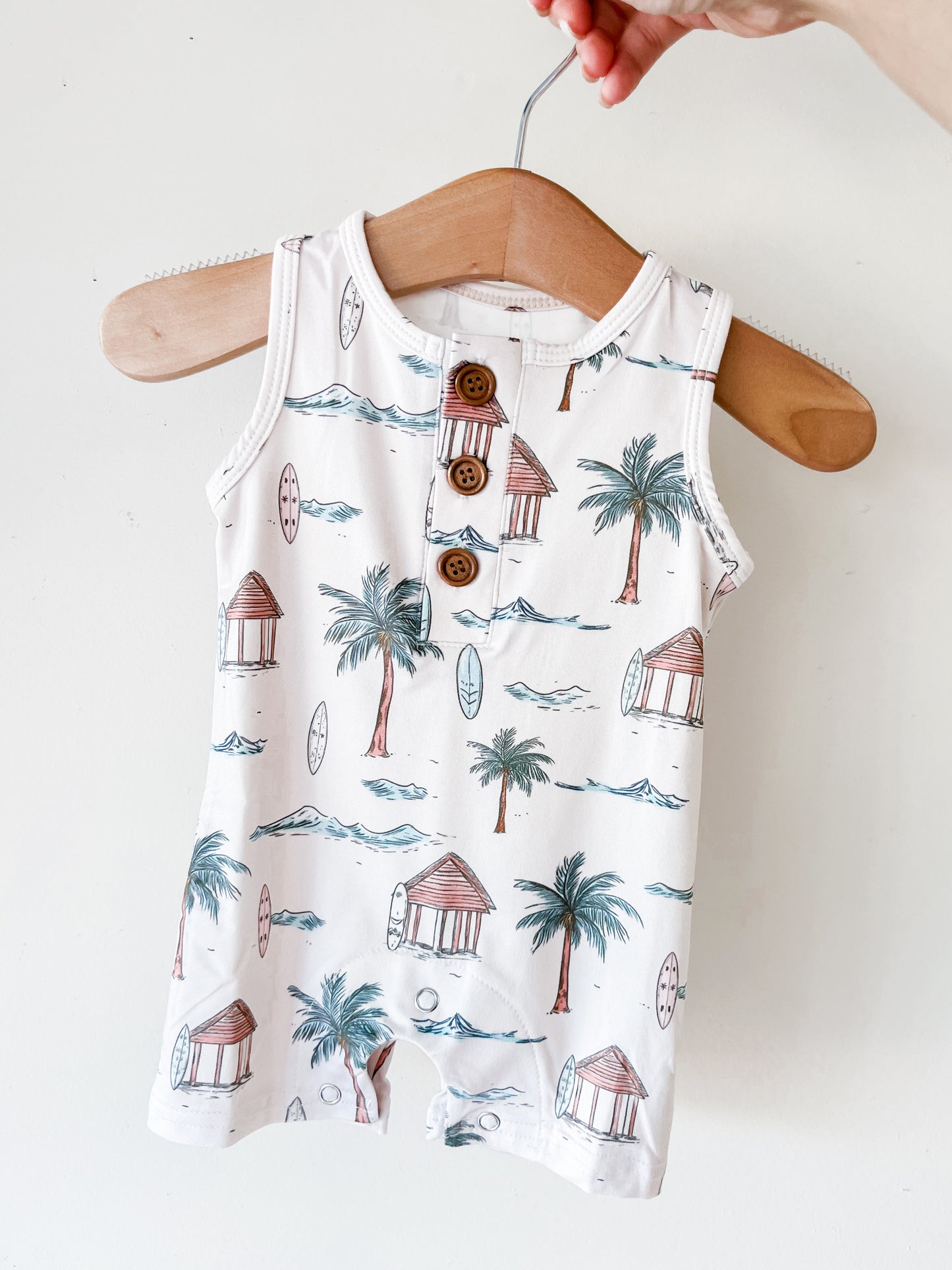 The Palm Coast Tank Romper by Tiny Tot Bundles is a sleeveless baby outfit with a tropical beach print—palm trees, waves, huts, and surfboards—shown on a wooden hanger held by hand against a white background.