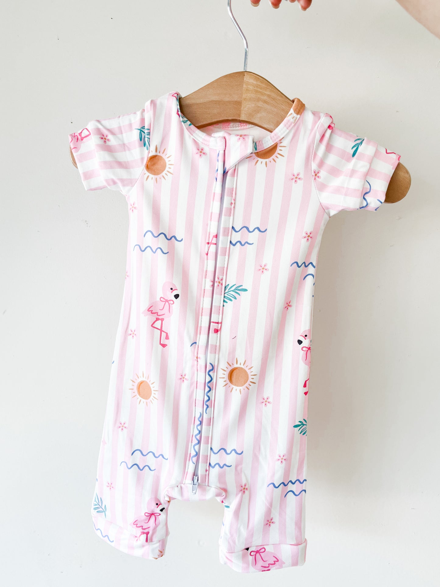 The Tiny Tot Bundles Pink Paradise Short Zippy, a short-sleeve romper with pink and white stripes featuring suns, waves, flamingos, and palm leaves, hangs on a wooden hanger against a white background.