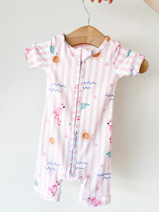The Tiny Tot Bundles Pink Paradise Short Zippy, a short-sleeve romper with pink and white stripes featuring suns, waves, flamingos, and palm leaves, hangs on a wooden hanger against a white background.