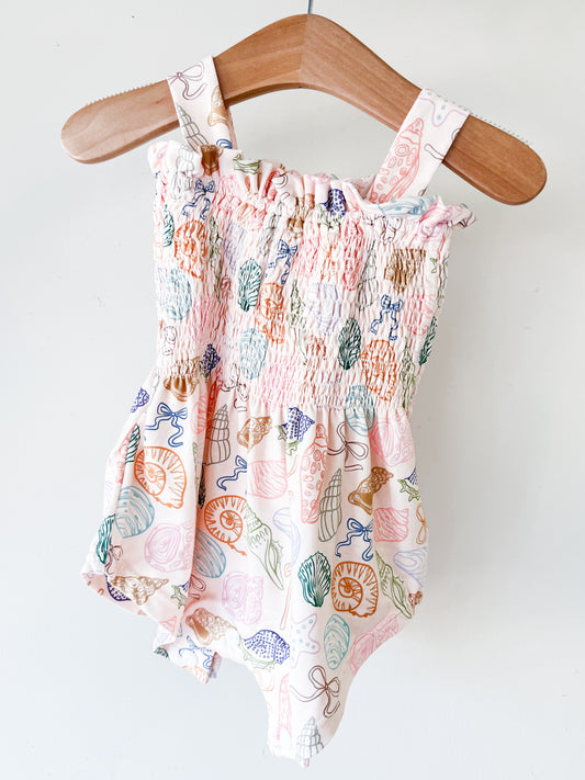 The Treasure Island Smocked Romper by Tiny Tot Bundles, featuring shoulder straps, a gathered elastic bodice, and a colorful seashell pattern, hangs on a wooden hanger against a white background.