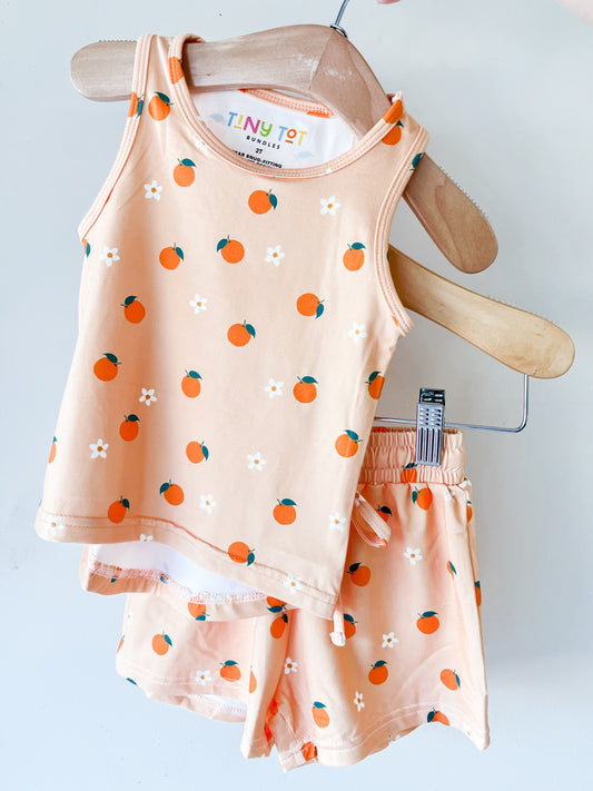 The Orange Blossom Tank Set by Tiny Tot Bundles features a peach toddler tank top and shorts with orange fruit and white flower prints, displayed on wooden hangers against a plain white background.